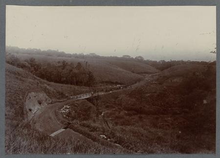 Construction of the road with bend and bridge through sloping terrain. Tipped-in photo in an album with 87 photos about the construction of the Gajoweg in North Sumatra between Bireuen and Takinguen between 1903-1914.